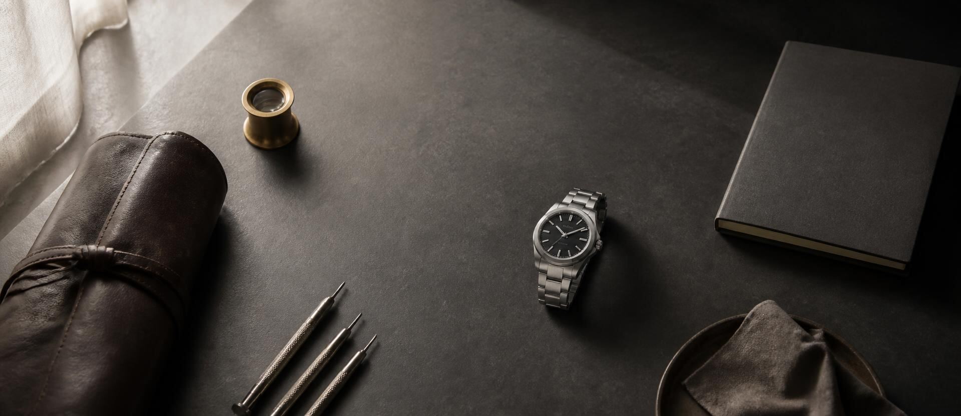 A specialist's desk with a leather watch roll, brass loupe, fine screwdrivers, a notebook and a single steel wristwatch